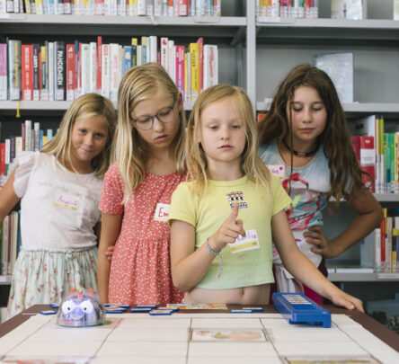 4 Mädchen stehen hinter einem Tisch, auf dem sich ein kleiner Roboter befindet